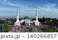 An aerial view of the Baitul Faizin Grand Mosque during the busy Eid al-Fitr prayer, with the signature green dome and twin minarets, a vast congregation on blue mats 140266857