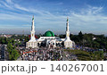 An aerial view of the Baitul Faizin Grand Mosque during the busy Eid al-Fitr prayer, with the signature green dome and twin minarets, a vast congregation on blue mats 140267001