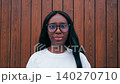 Smiling girl stands in front of wooden wall during bright day in urban area wearing glasses and white shirt 140270710