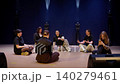 Group of young women sitting in a circle on a dimly lit stage, rehearsing and talking together in a 140279461
