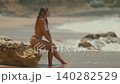 Woman enjoying sun at the beach while sitting on a rock beside the waves and ocean 140282529