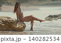 Woman sitting on rock at beach while waves lap at her feet in warm sunlight during summer 140282534