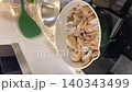 Asian female influencer cooking vlog video using smartphone to show spaghetti alle vongole recipe in kitchen with fresh clam parsley garlic green pot joyful atmosphere home interior modern utensil 140343499