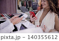 Smiling Woman Making Contactless Payment with Smartphone in Fashion Store. A cashier holds a POS terminal to pay for goods 140355612