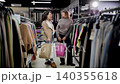 Brunette woman showing to female friend her purchase in paper bag. Young cheerful girls talking and making surprised facial expression while standing at mall after shopping 140355618