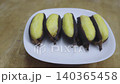 Thai grilled bananas in dark peels on a white plate over a wooden surface. Traditional healthy Asian street food snack or dessert. Close-up high angle shot of tropical fruit delicacies. 140365458