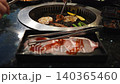 Raw sliced pork with red sauce in a black tray with tongs. In the background, meat and vegetables grill on a circular inset metal grate. Authentic Asian barbecue restaurant dining scene. 140365460