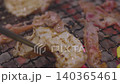 Close-up of thin marinated meat slices grilling on a wire mesh over hot burning charcoal. Traditional Japanese yakiniku or Korean barbecue style with red embers. Focus on texture and heat. 140365461