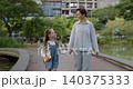 Portrait of little girl with schoolbag holds mother hand as walks along bridge over pond. Loving woman takes daughter to school across green urban garden 140375333