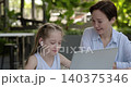 Portrait of happy little girl listens to video lesson via laptop as mother supports nearby in cafe. Woman sits near smart schoolgirl learning online in office 140375346