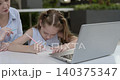 Portrait of little girl writes in notebook as mother sits nearby at laptop in office. Woman tutor teaches junior schoolgirl with online materials outdoors 140375347