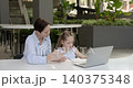 Portrait of attentive female teacher gives explanations to little girl at laptop in office. Mother checks daughter writing in notebook and looks at computer 140375348