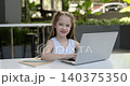 Portrait of pretty little girl with laptop and exercise book smiles at cafe table. Positive junior schoolgirl does homework online via computer on terrace 140375350