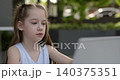 Portrait of dedicated little girl in blouse uses laptop for e-learning in outdoor cafe. Cute junior schoolgirl watches remote lesson via computer on terrace 140375351