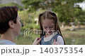 Portrait of little girl talks to mother as mom adjusts schoolbag in morning park. Loving woman helps junior schoolgirl to prepare for classes in urban garden 140375354