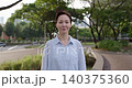 Portrait of middle aged woman with short haircut stands in green park. Positive lady wearing loose blue shirt smiles in summer urban garden. Urban lifestyle 140375360