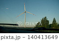 Car drives along rural road toward large wind turbine under clear blue sky. Scenic renewable energy landscape with trees on roadside. 140413649