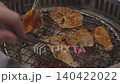 Slices of marinated meat grilling over red-hot charcoal on a wire rack. Metal tongs flip a piece of pork in a traditional Korean BBQ restaurant setting. Close up view of the cooking process. 140422022