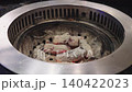 Close up of glowing charcoal embers in a round metal tabletop grill at a restaurant. Metal tongs adjusting burning coals for Japanese yakiniku or Korean barbecue cooking. Intense heat. High angle. 140422023