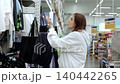 Young woman browsing through colorful clothing in a lively store, holding a navy blue garment on a hanger. Bright displays and diverse styles create an inviting shopping vibe 140442265