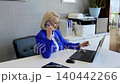 Businesswoman in a blue suit sits at her desk, talking on her phone and working on her laptop. She is focused and efficient, multitasking with ease 140442266