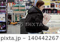 Young woman in a black winter coat shops for frozen food. Opening the sliding glass door of a chest freezer, she selects various items to purchase 140442267