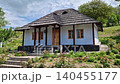 Traditional Moldovan peasant house with burned shingle roof. The cottage features white clay-plastered walls, wooden windows, and a porch supported by dark timber pillars, fronted by a blooming garden 140455177