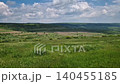 A vast green meadow rolls across a scenic Moldovan valley under a bright, cloudy sky. Sprawling agricultural fields, dense distant forests, and green hillsides. Rural landscape of patchwork plains 140455185