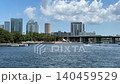 Skyscrapers and urban buildings stand behind a lush green shoreline along a sparkling waterway with boats. A large bridge spans the river under a bright blue sky filled with white puffy clouds. 140459529