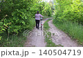 Caucasian woman in plaid shirt walking along a dirt road pushing a bicycle surrounded by lush green summer forest, concept of active recreation and healthy lifestyle 140504719