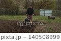 Male farmer using a motor cultivator to till the ground in his backyard, preparing the soil for planting seeds during the spring agricultural season with specialized farming equipment 140508999