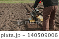 Male farmer using a garden rototiller to plow the soil and prepare the land for planting spring crops, turning up fresh dark earth in neat rows on a sunny day in the backyard 140509006