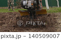 Powerful walk-behind tractor blades digging into the ground, cultivating soil for planting in a garden or farm field while a farmer guides the agricultural machinery from behind. 140509007