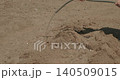 Close-up shot of a female gardener's hands watering small holes in a dry, sandy garden bed with a hose, meticulously preparing the soil for planting young vegetable seedlings in spring. 140509015