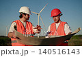 Engineer and worker in hard hats and orange safety vests review large construction plan at wind farm site. Man points to blueprint while colleague studies project documentation with wind turbines in 140516143