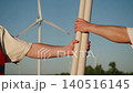 Construction engineer in white hard hat hands over technical documentation to field worker during renewable energy project. Close up of hand exchange, orange safety vest, and wind turbine blades in 140516145