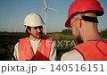 Engineer in white hard hat reprimands worker in red helmet at wind farm site. Boss points at documentation while explaining mistakes near wind turbines in rural field 140516151