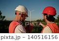 Two industrial workers in protective helmets and high visibility vests pause work at renewable energy site. Cigarettes in hands and wind turbines in background show casual break during project 140516156