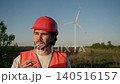 Wind farm technician wearing protective helmet and reflective vest takes smoking break outdoors. Man holds cigarette near face while wind turbines and countryside landscape remain blurred in 140516157