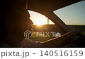 Driver seen in soft focus drives car at sunset with warm sunlight shining through window. Man appears to speak while traveling through rural landscape with forest visible in background during evening 140516159