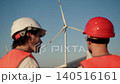 Engineers communicate while reviewing wind turbine documentation at wind farm site. One engineer in white hard hat and other in red helmet discuss project details with wind turbine visible in 140516161