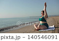 Caucasian woman on seashore on towel does warm up, stretches arms up and bends. Morning meditative exercises of person on beach and enjoying view. Self care and inspiration on vacation. 140546091