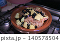 Close up view of covering a traditional moroccan tagine with raw chicken thighs, diced potatoes, and black olives, ready for slow cooking on a gas stove in a cozy home kitchen setting 140578504