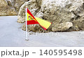 Small Spanish flag planted in sandy beach near large rocks, with the flag waving gently in the breeze, showcasing vibrant colors of red and yellow against a natural coastal backdrop 140595498