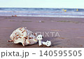 Shell on sandy beach with wireless earbuds positioned beside it, capturing the serene coastal atmosphere with gentle waves and distant beachgoers visible in the background 140595505