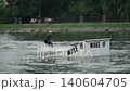 A male athlete black wetsuit yellow helmet holds onto rope one hand and trains wakeboard. city lake, people strolling along embankment background, ramp foreground, a cloudy summer day. 140604705