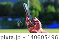Male hand scrolling content on smartphone near football stadium field at sunset, close up of man using mobile phone browser outdoors, social media browsing, mobile internet, online communication 140605496