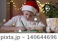Boy with cast on hand and Santa hat writing letter to wizard at home with focused expression, arc gimbal shot. Child writing wishes and telling about desired gifts in decorated room with warm lights. 140644696