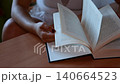 Unrecognizable young woman sitting at a wooden table and slowly turning the pages of a book, representing a moment of quiet relaxation, education, and enjoying literature at home 140664523