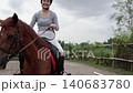 Woman riding chestnut horse along rural path from start to finish under cloudy sky 140683780